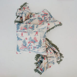 A square throw pillow and matching fringed throw blanket featuring a vintage winter fairytale scene print in red, blue, and green on a cream background, displayed on a textured white wall.