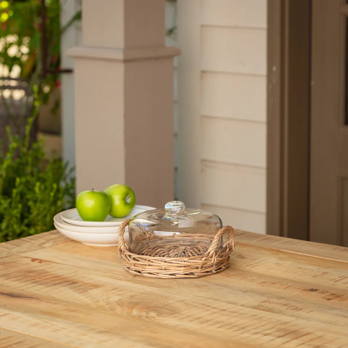 Clear glass cloche lid on a woven basket tray with green apples on a plate outdoors.