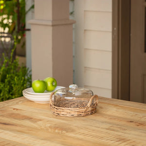 Clear glass cloche lid on a woven basket tray with green apples on a plate outdoors.