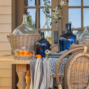 Outdoor table setting with blue bottles, wicker basket, and oranges near a window.