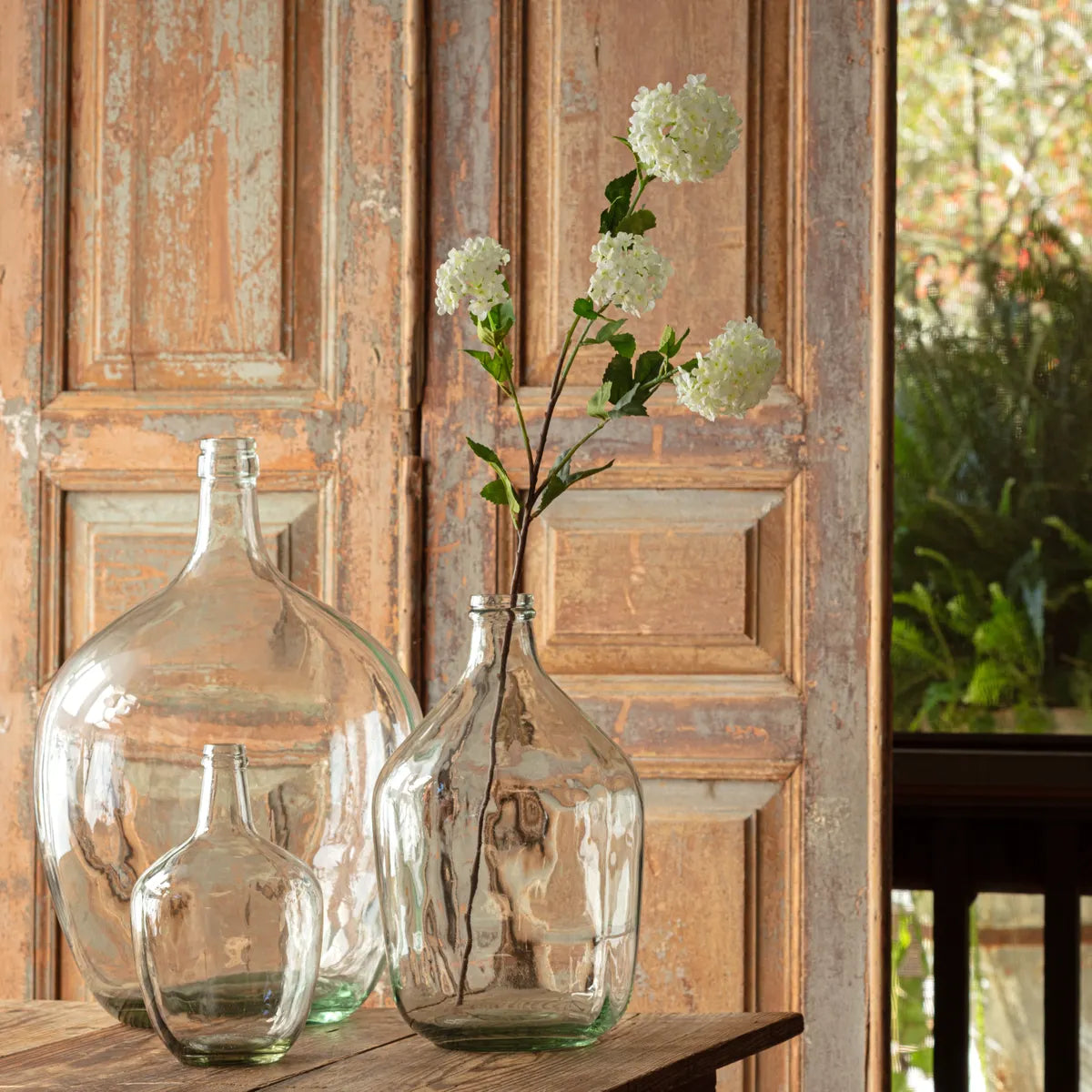 Clear sea glass bottles with white snowball viburnum faux floral stems on a wooden surface with a rustic wooden door in the background.
