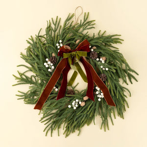 Christmas wreath with greenery, berries, pinecones, and multicolored velvet ribbons on a beige background.