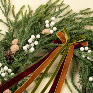 A close-up of a festive green wreath made of pine branches, decorated with white berry clusters, small pinecones, and a large velvet ribbon bow in rust brown, olive green, and burgundy on a cream background.