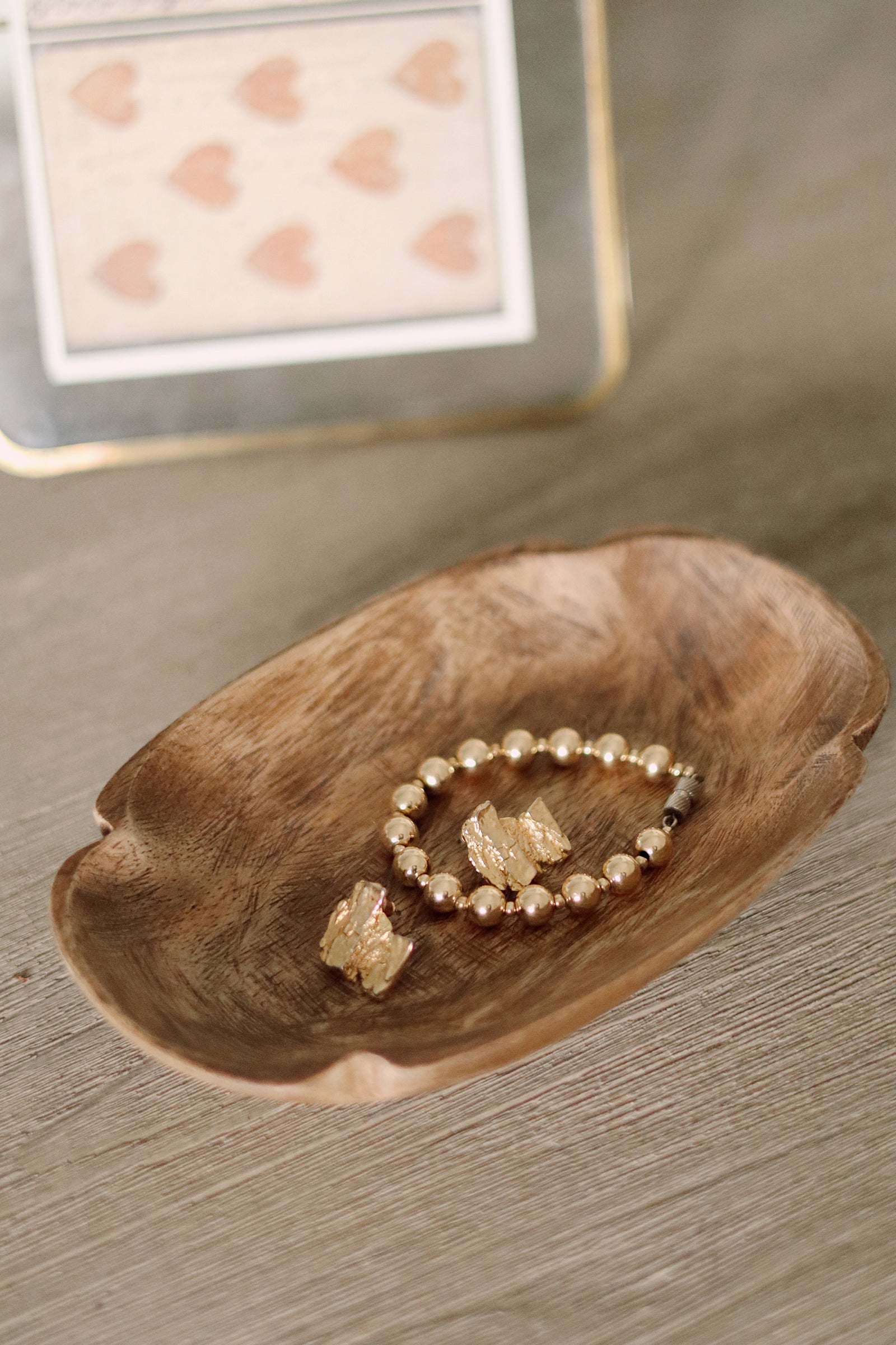 A close-up of a wooden bowl containing a beaded bracelet and a small crystal charm, placed on a gray wood-grain surface with a blurred decorative card in the background.