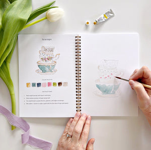 Person drawing spring tea cups in a watercolor sketchbook with a tulip and paint tubes on a white surface. Emily Lex Studios. 