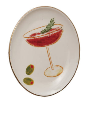 An oval ceramic plate with a hand-painted illustration of a red cocktail in a coupe glass, garnished with a green herb sprig, and two green olives with red pimentos. The plate has a gold rim and is set against a plain white background.