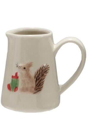 An off white ceramic creamer pitcher featuring a hand-painted illustration of a brown squirrel holding a red and green stocking, set against a plain white background.