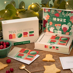 A boxed holiday card game set labeled 'TELL ME MORE HOLIDAY EDITION' arranged on a wooden table with festive props. The white product box and inner packaging feature colorful 'MORE' lettering and illustrated holiday-themed graphics.