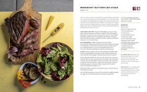 Steak on a cutting board with a side of salad and vegetables on a yellow surface, next to a magazine article. Recipe from inside book "The Meateater Outdoor Cookbook" Steven Rinella. 