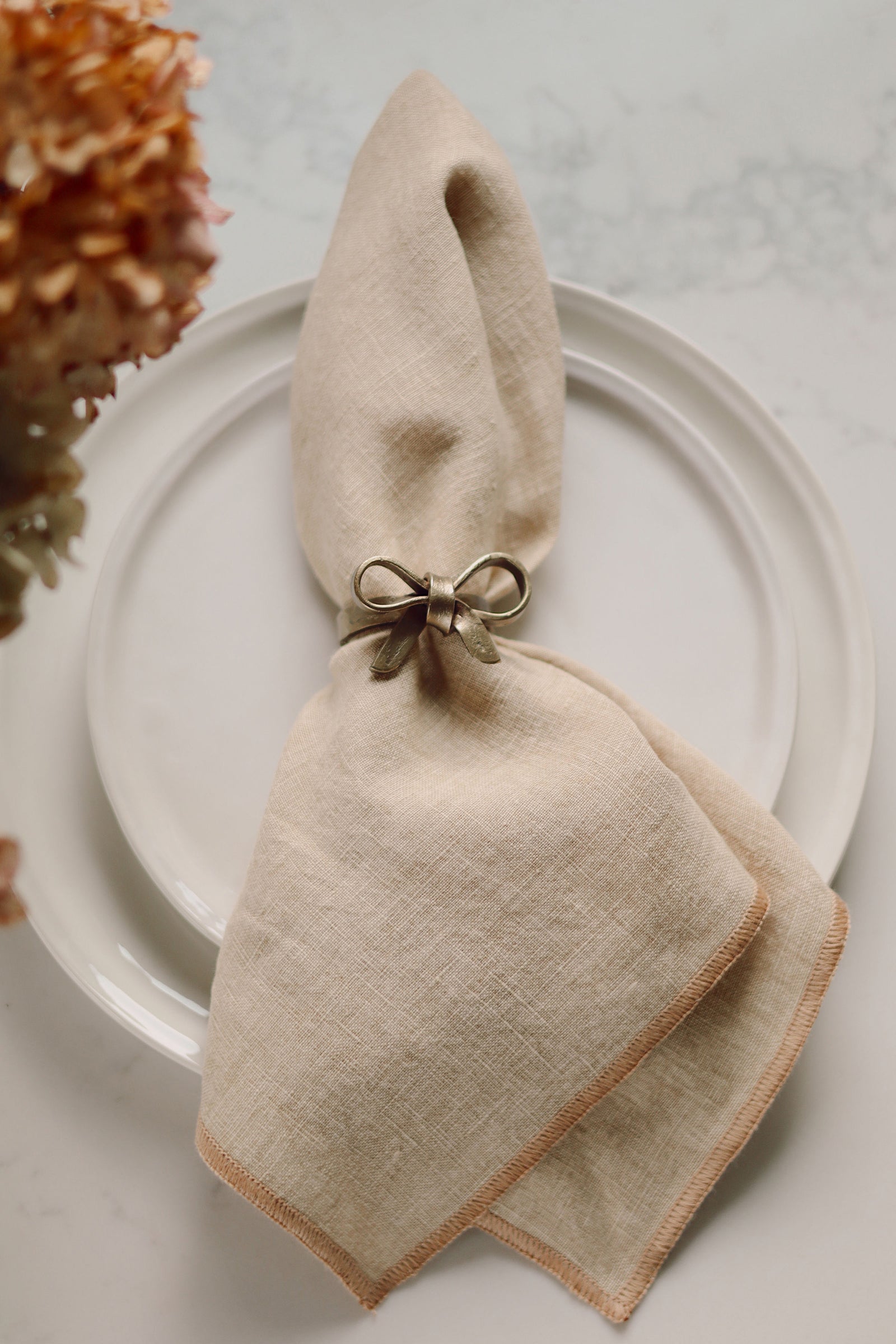 A beige linen napkin folded and tied with a metallic gold iron bow napkin ring, presented on a white plate with dried flowers in the corner.