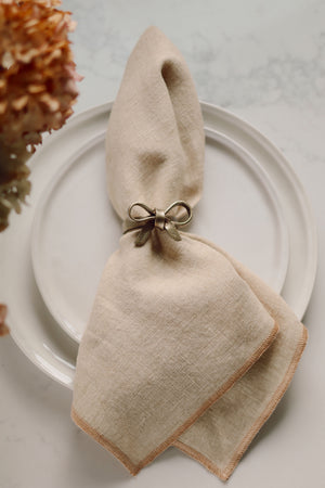 A beige linen napkin folded and tied with a metallic gold iron bow napkin ring, presented on a white plate with dried flowers in the corner.