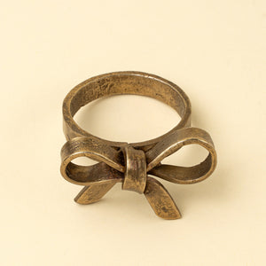 A vintage-style bronze tied gold bow napkin ring, shown on a plain beige background.