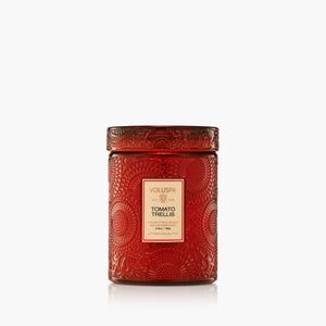 A red textured glass jar candle with a matching lid, featuring a beige label that reads 'VOLUSPA TOMATO TRELLIS'. The candle is photographed on a clean white background with a subtle reflection underneath.