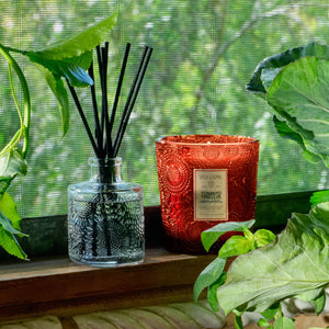 A reed diffuser bottle with black reeds and a textured red-orange candle jar labeled Tomato Trellis sitting on a wooden windowsill with green leaves framing the scene.