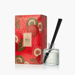 A reed diffuser bottle with black reeds next to its red patterned packaging box, featuring the brand name Voluspa and the new scent  "TOMATO TRELLIS"