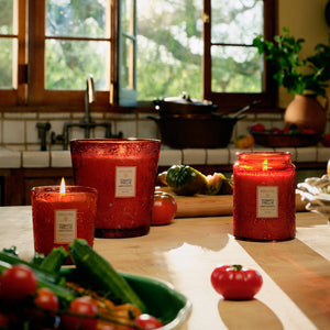 Red glass Tomato Trellis candles in various sizes on a kitchen counter with vegetables and a window in the background. Voluspa. 