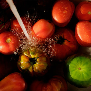 Fresh tomatoes being washed under running water, with red Roma-style tomatoes, a green heirloom tomato, and a yellow-green tomato visible against a dark background.