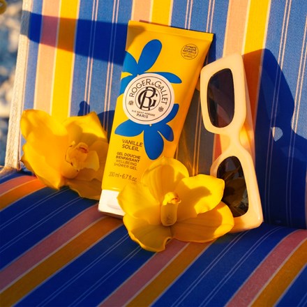 A tube of Roger&Caviar Vanille Scher body cream sits on a blue, yellow, and orange striped beach chair, styled with yellow daffodils and white sunglasses under bright sunlight.