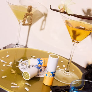 Two cocktail martinis glasses filled with amber liquid, styled on a gold tray with lip balm tubes and confetti. The background is a warm beige wall with soft shadows.