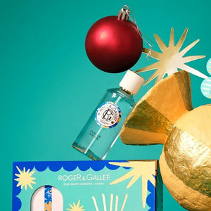 A Roger & Gallet Vanille Soleil Eau de Toilette bottle with a turquoise blue liquid and white cap, styled with a red Christmas ornament, gold star cutouts, and a gold bow against a teal background.