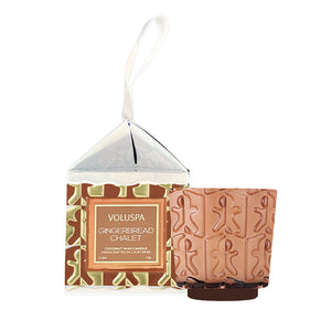 Voluspa Gingerbread Chalet scented wax candle in a clear hanging gift box with a brown and gold patterned label, shown next to a small gingerbread-themed candle holder in a warm beige-brown tone on a white background.