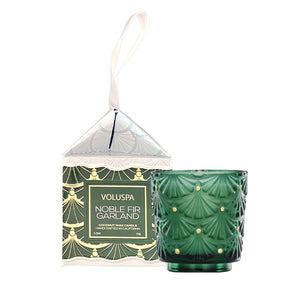 Voluspa Noble Fir Garland coconut wax candle in a dark green textured glass holder, shown next to its illustrated window box packaging with a white hanging ribbon.