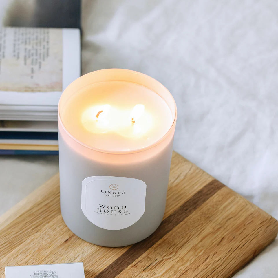 Lit candle in a glass container labeled 'Wood House' on a wooden surface with a blurred background.