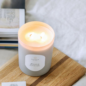 Lit candle in a glass container labeled 'Wood House' on a wooden surface with a blurred background.