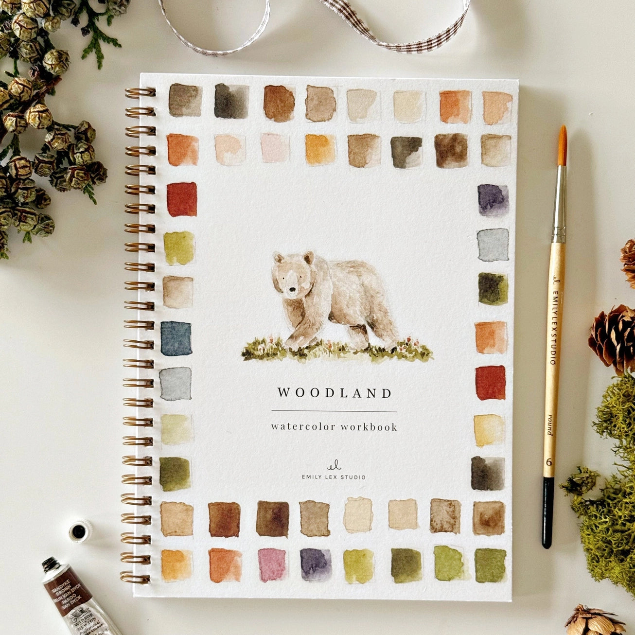 Woodland watercolor workbook with a bear illustration on a white surface with decorative woodland elements. Emily Lex Studios. 