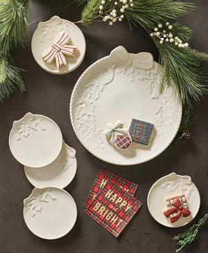 A set of white ceramic holiday plates with embossed floral and bow motifs, arranged on a dark surface with green pine branches and white berries. Decorated cookies and a red plaid napkin with 'HAPPY BRIGHT' are styled around the plates.