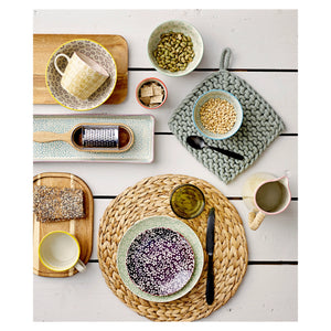 Display of various colors, patterns, and textural kitchen utensils including: acacia wood cheese grater, mugs, plates, bowls filled with beans and crackers, and pot holders. 