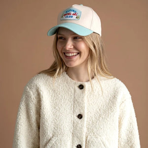 A smiling blonde woman wearing a cream sherpa fleece button-front cardigan and a cream baseball cap with a light blue visor. The cap features a front patch that reads 'ALPINE SKI CLUB'.