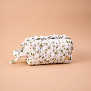A quilted cosmetic pouch with a white background and a green pine tree pattern, featuring a ruffled top edge, a matching fabric handle, and a small gold logo patch on a solid peach background.