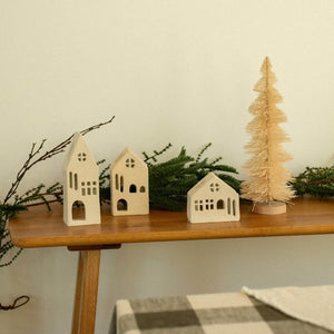 Three small white metal house ornaments and a matching beige bottle-tree decoration displayed on a wooden shelf with greenery against a light wall.