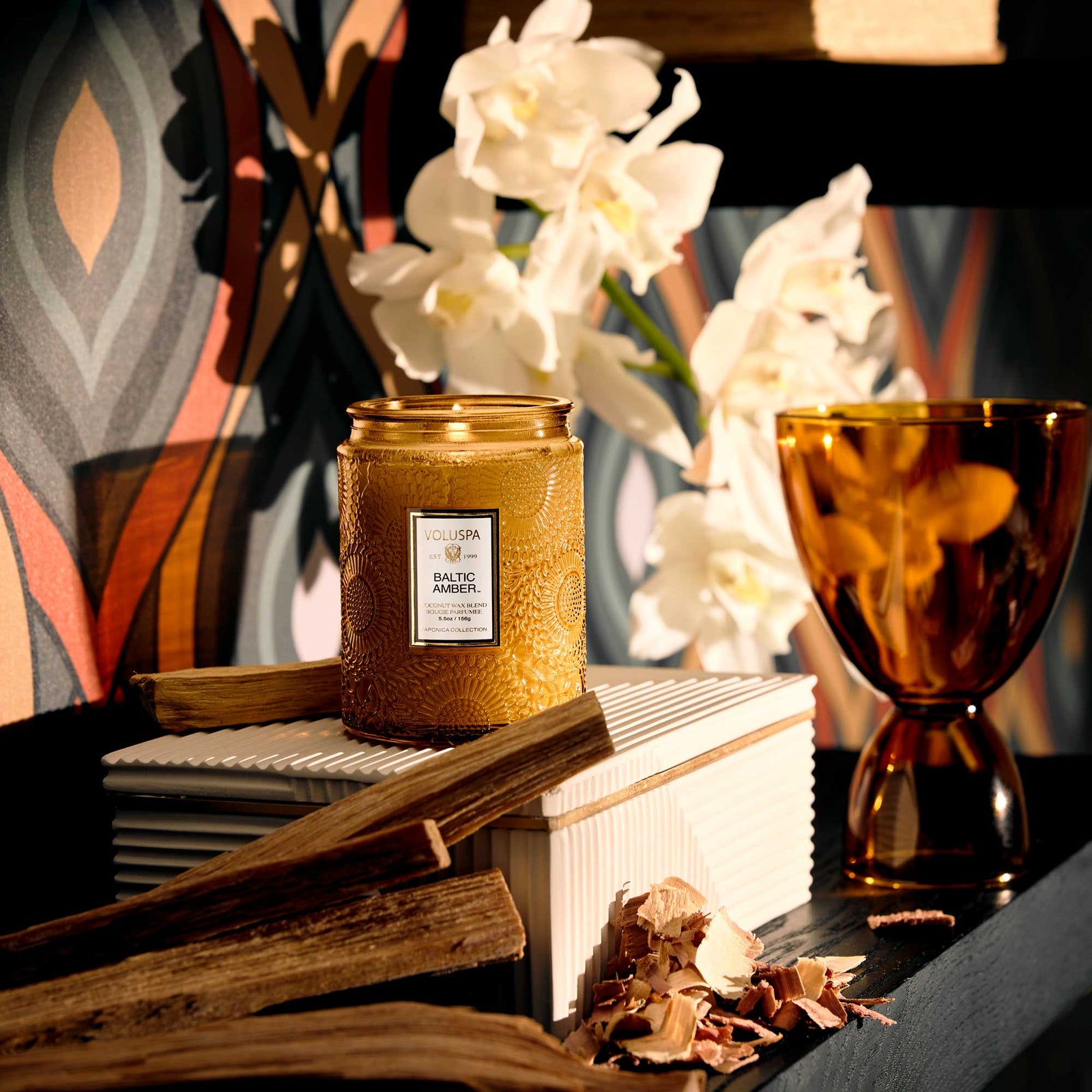 Decorative display with Baltic Amber candle, books, and white orchid flowers on a textured console table. Voluspa. 