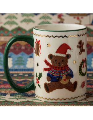 A white ceramic Christmas mug with a dark green handle and rim, decorated with a teddy bear wearing a red Santa hat and multicolor knit sweater, surrounded by holiday motifs like a wreath, gingerbread man, and bells, set against a patterned background.