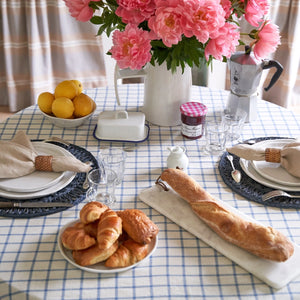 Set table with croissants, baguette, and pink flowers on a blue windowpane tablecloth.