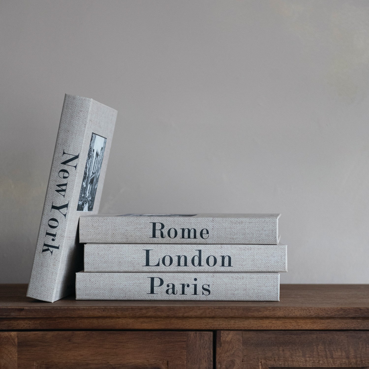 Stack of books titled 'New York', 'Rome', 'London', and 'Paris' on a wooden surface with a neutral background.