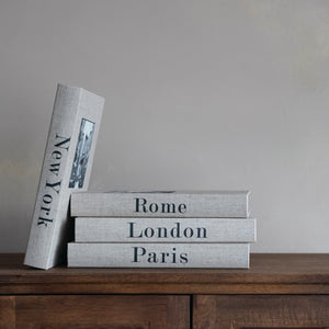 Stack of books titled 'New York', 'Rome', 'London', and 'Paris' on a wooden surface with a neutral background.