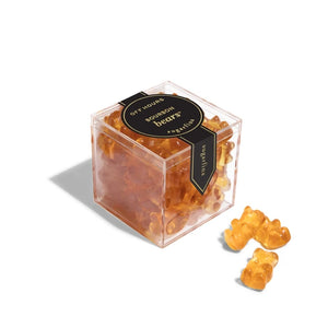 A clear acrylic cube container filled with amber-colored gummy bears, with two gummy bears placed outside the box on a white background. The box has a black label reading 'OFF HOURS Bourbon bears Sugarfina'.