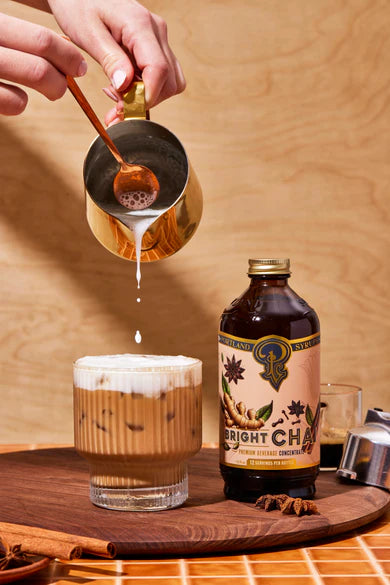 Person pouring a chai latte into a ribbed glass with a bottle of 'Bright Chai' on a wooden surface. Portland Syrups. 