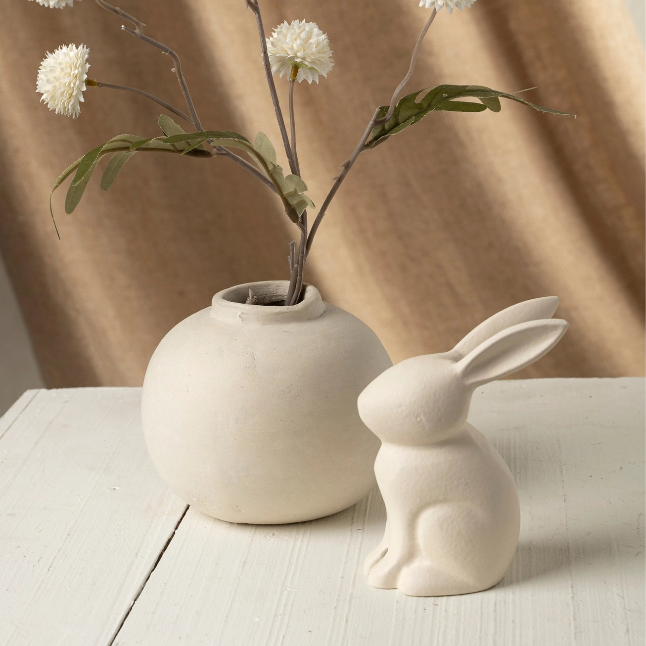 Ivory ceramic vase with flowers and a matching ivory rabbit figurine on a wooden surface with a beige curtain background.