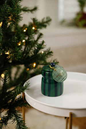 A small green striped jar with a matching lid and a round tag, styled on a white tray next to a Christmas tree with warm lights.
