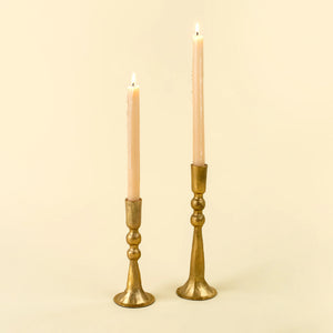 Two lit taper candles in ornate gold-toned metal candlesticks against a warm cream background.