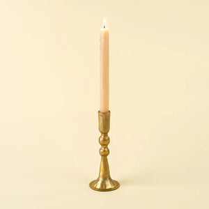 A single lit taper candle in a slim brass candlestick holder against a plain cream background.