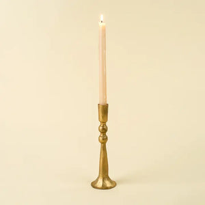 A single lit taper candle in a tall, flared brass or gold-toned holder against a plain warm cream background.