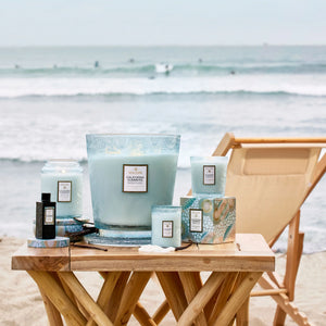 Various sized blue glass jar "California Summers" candles on a teak wood table set against the ocean. Voluspa. 