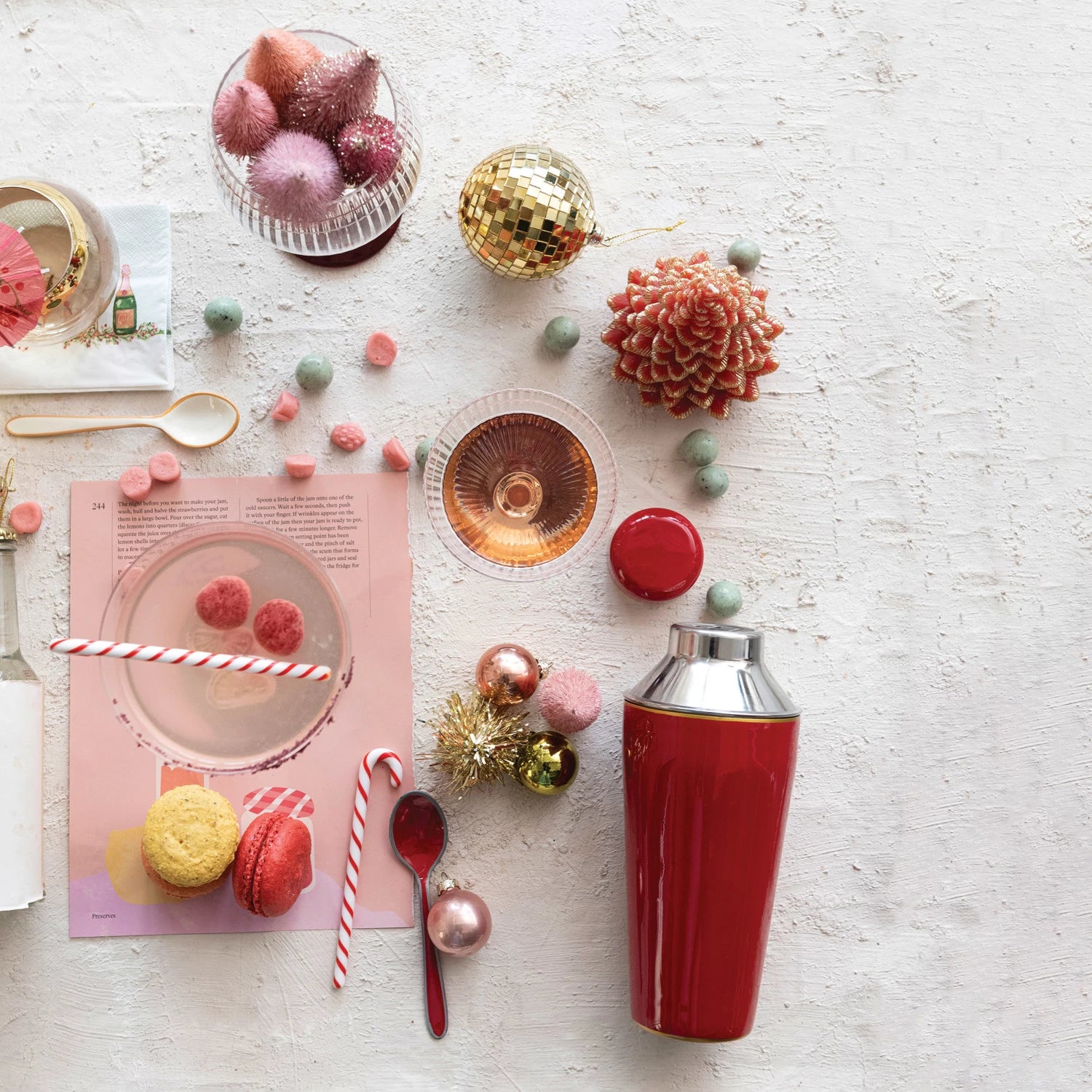 Decorative holiday table setting with a red martini shaker, candy canes stir sticks, and Christmas ornaments on a light surface.