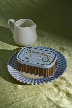 A rectangular metal candle tin labeled Cappuccino sits on a blue-and-white patterned plate, with a small white pitcher behind it on a green fabric surface.