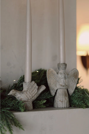 A close-up of a holiday mantel or shelf display featuring two white taper candles in rustic stone angel and dove-shaped holders, surrounded by green pine garland.